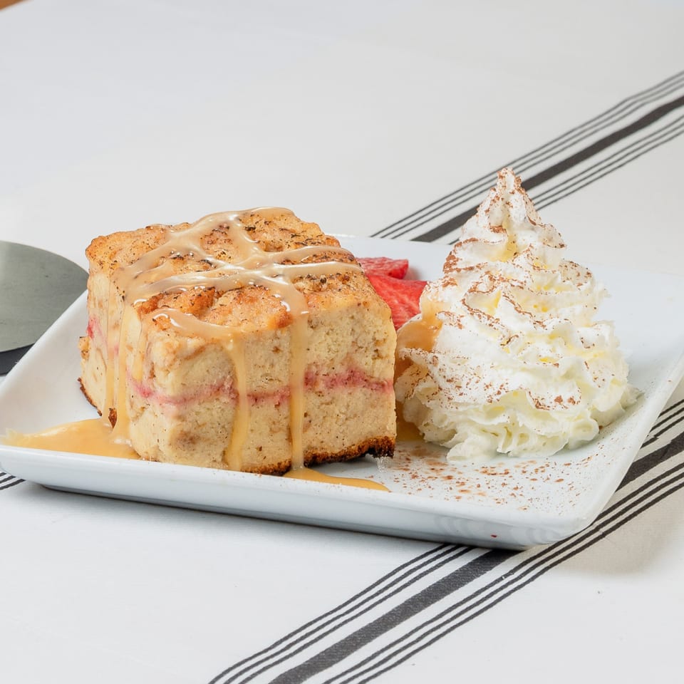 A slice of bread pudding with caramel drizzle, strawberries, and whipped cream on a white plate.