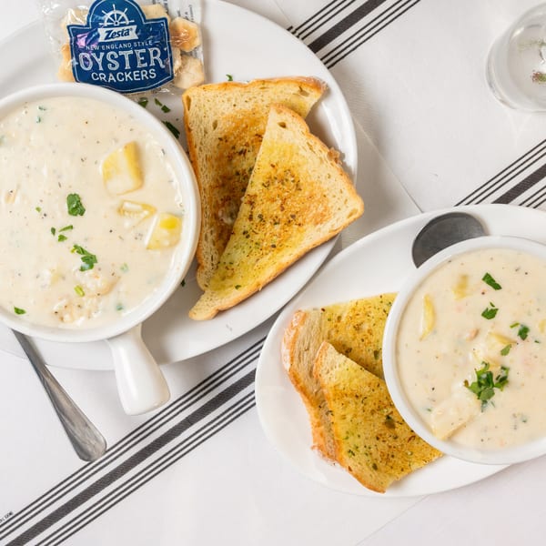 Two bowls of creamy clam chowder with garlic toast and oyster crackers on a striped tablecloth.