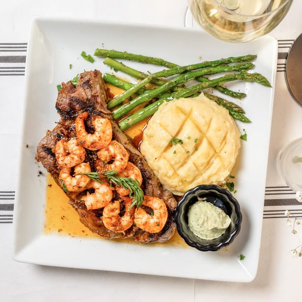 Grilled steak and shrimp with mashed potatoes, asparagus, and a side of sauce on a white plate.