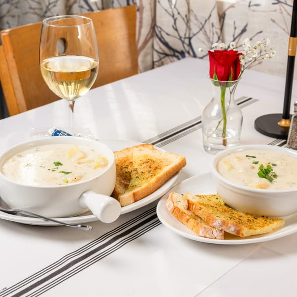 Two bowls of creamy clam chowder with garlic bread and white wine on a white tablecloth.