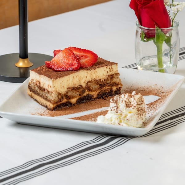 A slice of tiramisu topped with strawberries and cocoa powder, with whipped cream on a white plate.