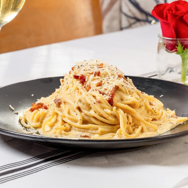 Creamy spaghetti carbonara with bacon bits and grated cheese on a black plate, with a red rose.