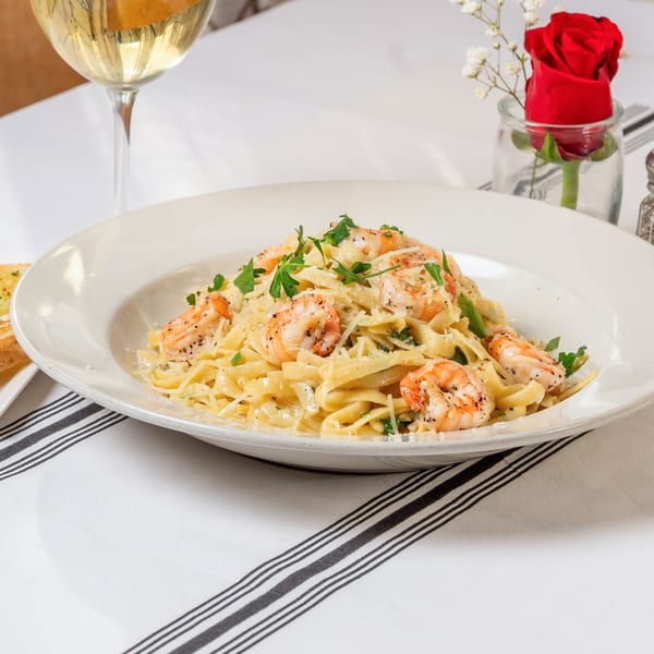 Shrimp fettuccine alfredo with parsley and parmesan, white wine, and a red rose on a striped tablecloth.