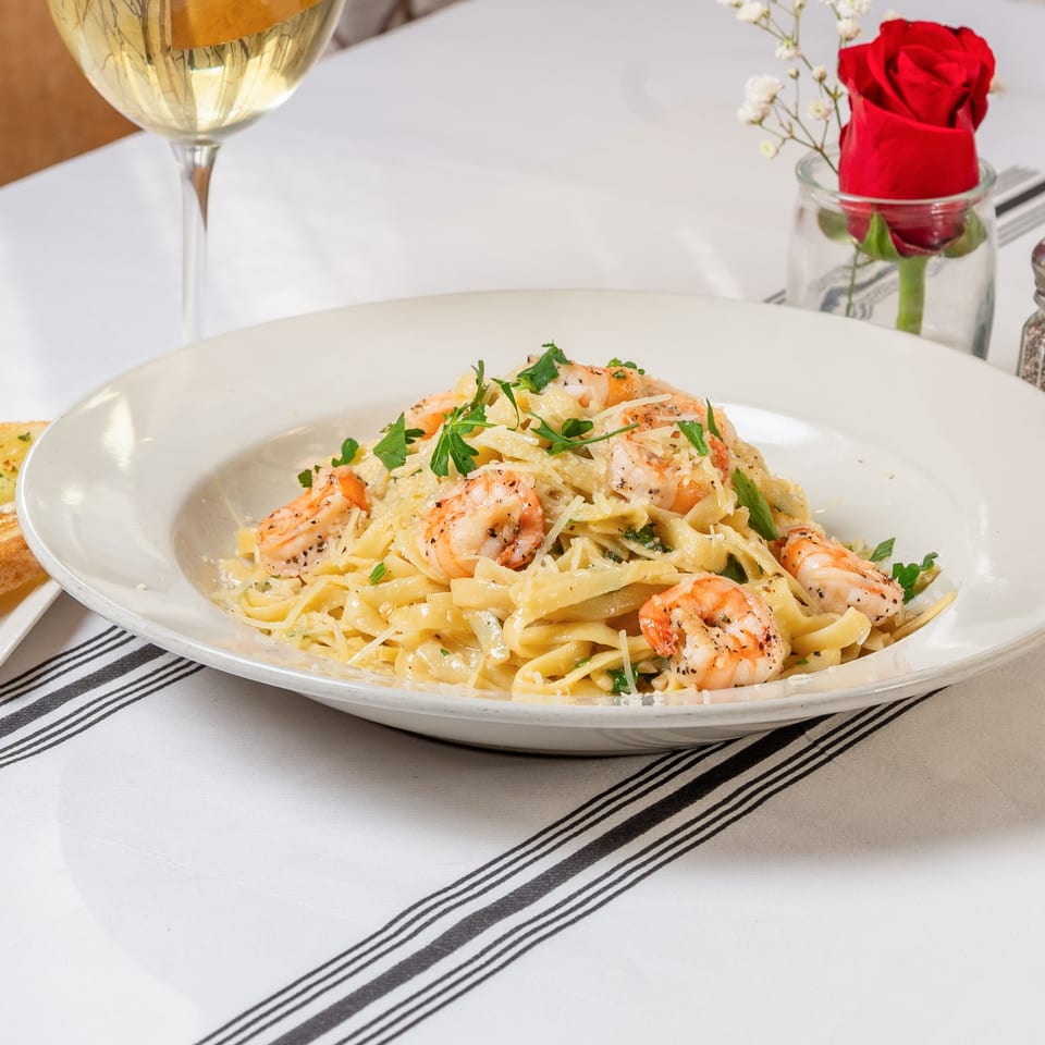 Shrimp fettuccine alfredo with parsley and parmesan, white wine, and a red rose on a striped tablecloth.